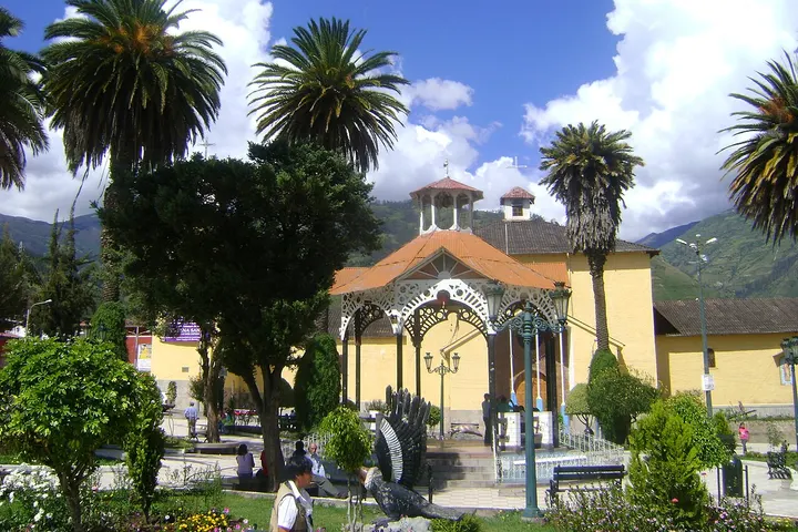 En primer plano, una fuente de agua con un arco de piedra, rodeada de palmeras y árboles