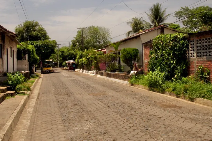 En primer plano, una calle pavimentada con adoquines, rodeada de casas y árboles