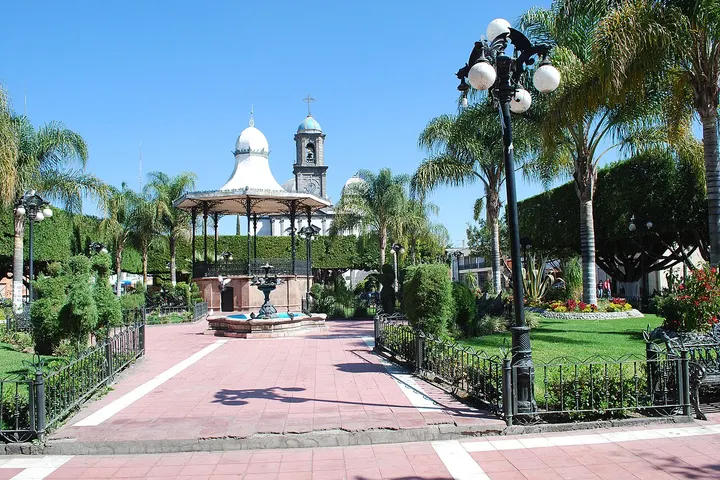 En primer plano, un parque con un kiosco y una fuente, rodeado de palmeras y un jardín