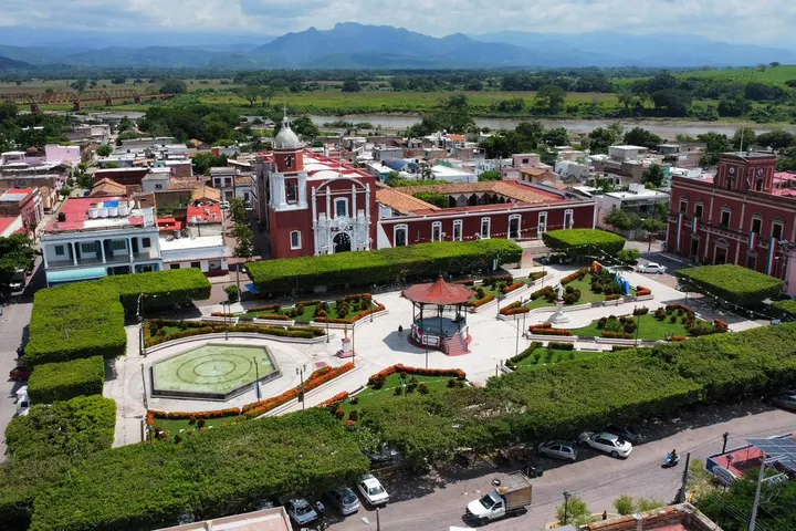 En primer plano, un parque con un kiosco y un jardín de flores, rodeado por un muro de hierba