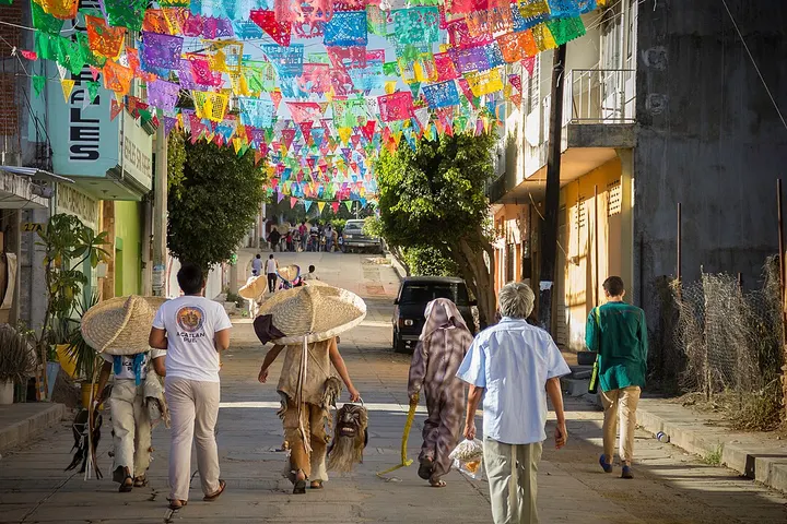 En primer plano, se ven personas caminando por una calle con una decoración de papel picado sobre sus cabezas