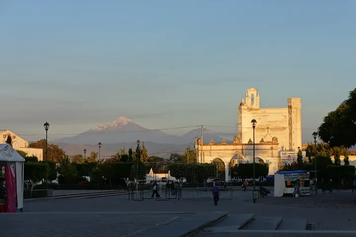 En primer plano, una plaza con una iglesia blanca y un monte nevado en el fondo