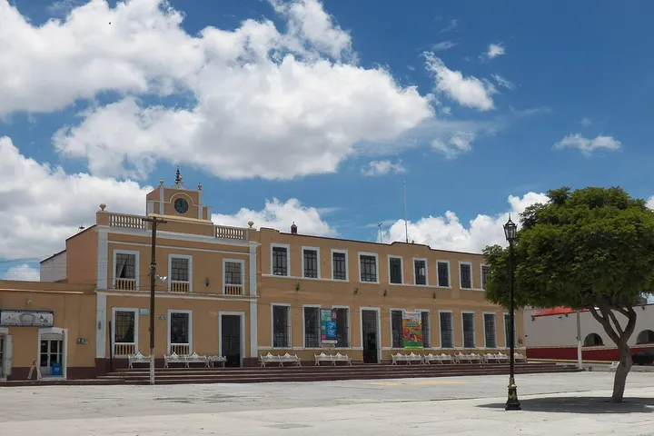 En primer plano, una gran casa de dos pisos, con balcones y una torre, rodeada por árboles y una plaza