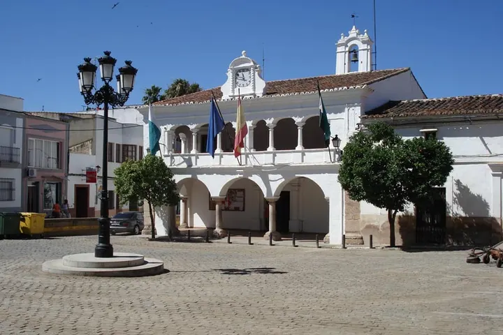En primer plano, una plaza con un farol de gas y árboles