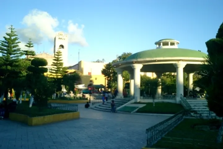 En primer plano, una plaza con un kiosco de estilo clásico, rodeada de árboles y una iglesia en el fondo