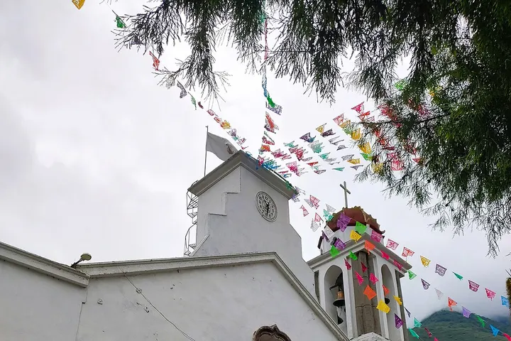 En primer plano, una iglesia blanca con un reloj y una cruz, con banderas de colores colgando del tejado