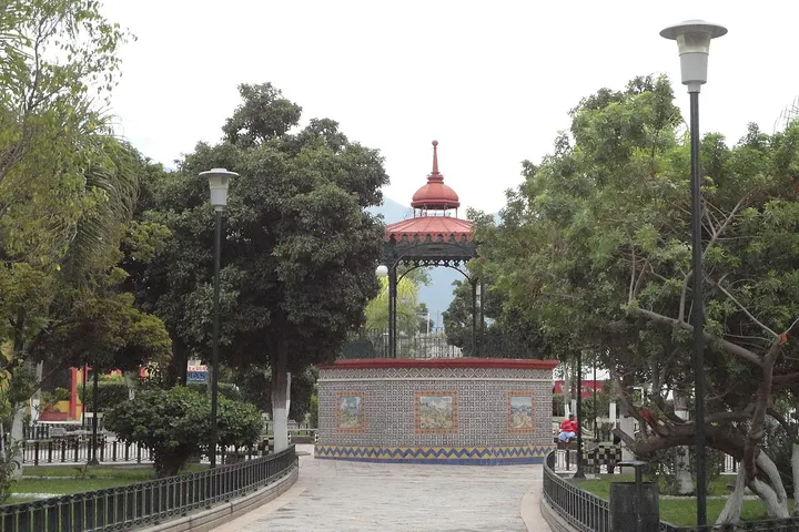 En primer plano, un jardín con un paseo y un monumento de estilo mexicano con un reloj de sol
