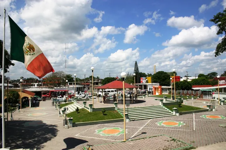 La bandera de México ondea en primer plano, en medio de una plaza con escalinatas y un pequeño parque