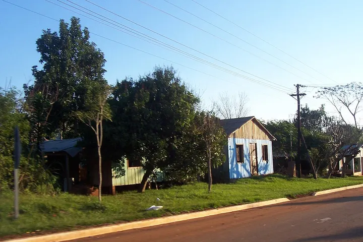 En primer plano, una casa con una fachada azul y un techo de madera, rodeada por árboles y una calle