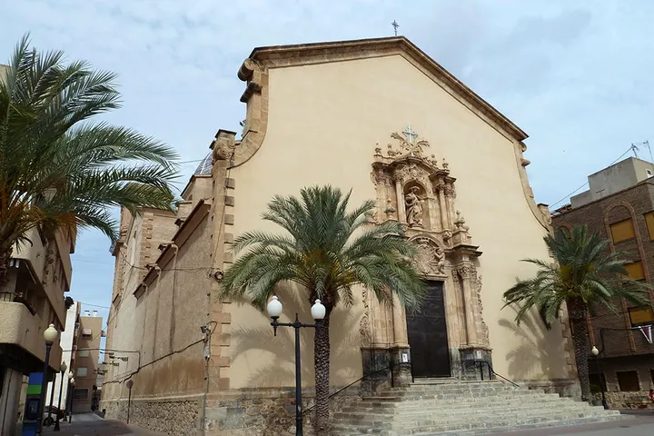 En primer plano, una iglesia con un gran portal y dos palmeras