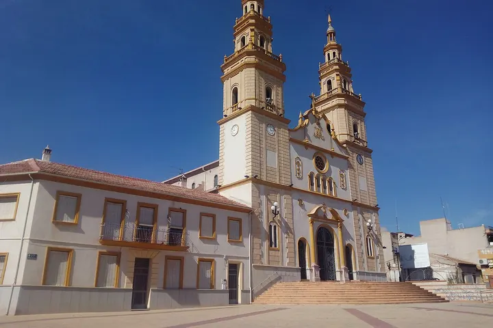 En primer plano, una iglesia con dos torres y un reloj en su fachada