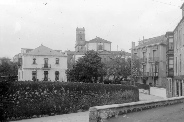 En primer plano, un muro de piedra con un seto de arbustos, y detrás, una iglesia y edificios de piedra