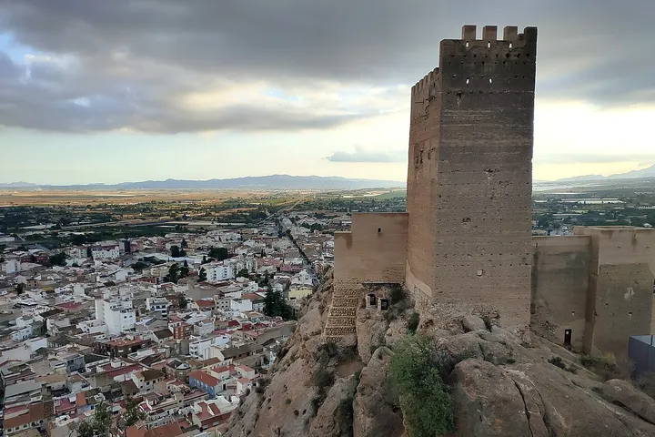 En primer plano, una torre de piedra con una vista panorámica de una ciudad con casas blancas y tejados rojos