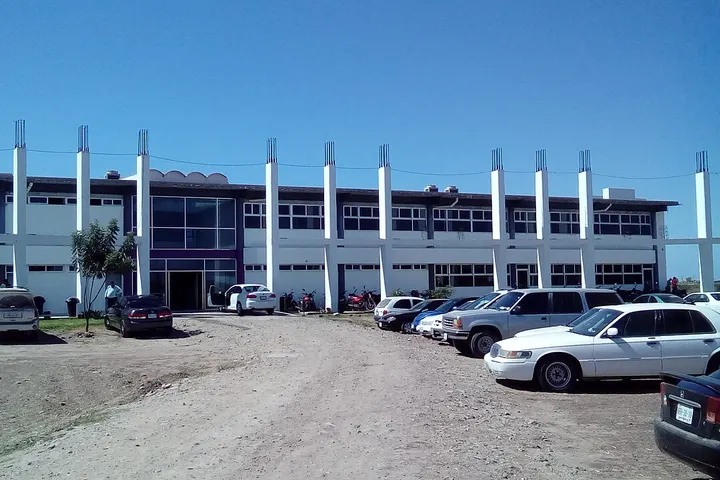 En primer plano, un edificio blanco con columnas blancas y un parqueo con coches