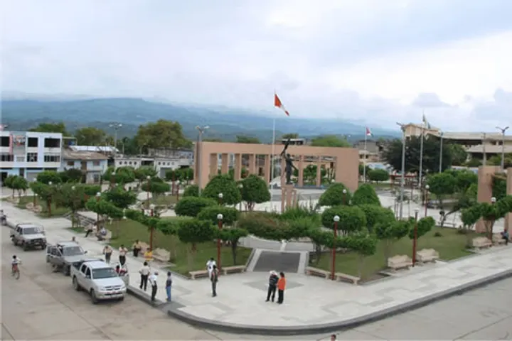 En primer plano, se ve una plaza con árboles y un monumento en el centro
