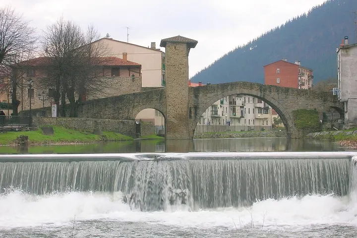 En primer plano, un impresionante salto de agua, con una antigua puente de piedra detrás