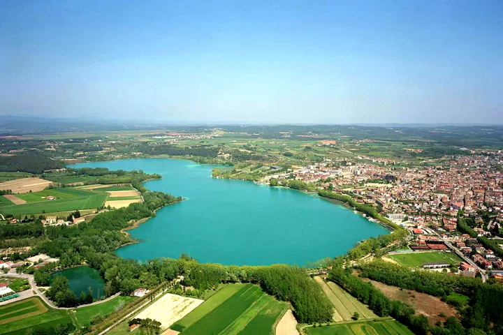 En primer plano, un lago rodeado de árboles y campos de cultivo