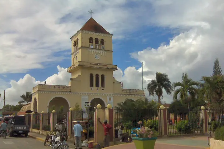 En primer plano, una iglesia con un campanario y una torre, con una entrada arqueada y un jardín con palmeras
