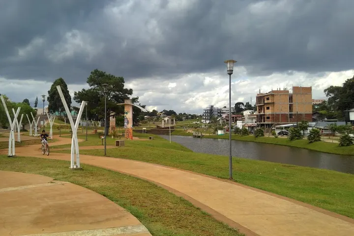 En primer plano, se ve un parque con un sendero, árboles blancos, una persona caminando y un río