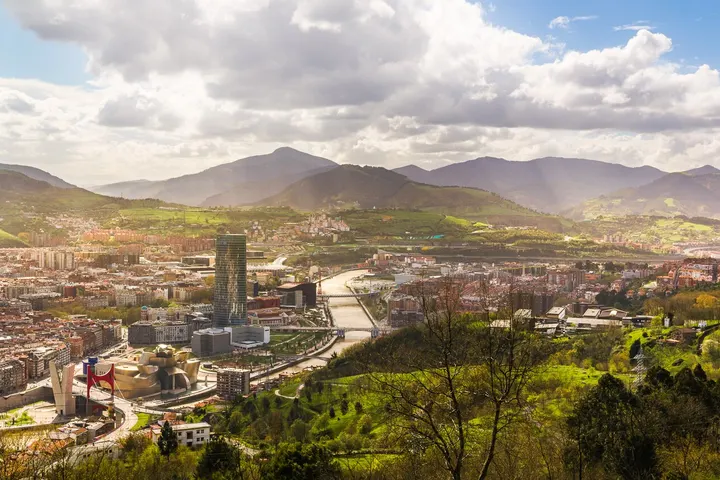 Una ciudad con montañas en el fondo, un río, un puente, edificios, calles, árboles y un cielo con nubes