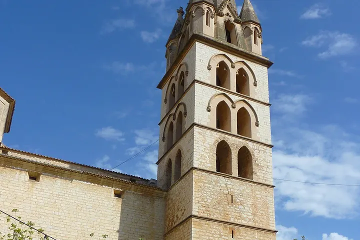 En primer plano, una torre de iglesia con arcos y pináculos, con un cielo azul y nubes blancas