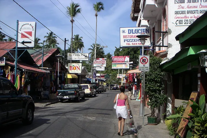 En primer plano, una mujer camina por la calle, rodeada de tiendas y edificios con letreros en un idioma desconocido