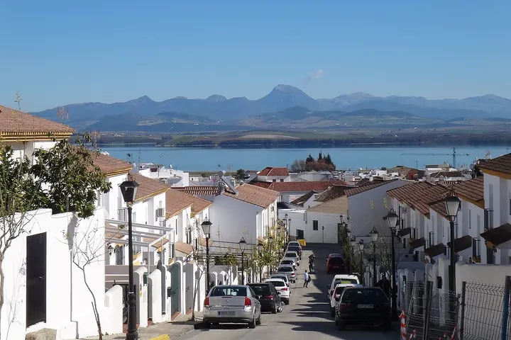 En primer plano, una calle con casas blancas y tejados rojos, con un lago y montañas en el fondo