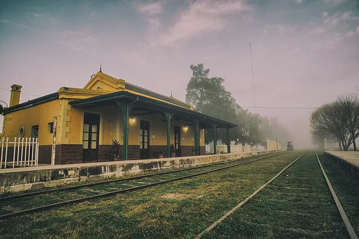 Un edificio amarillo con un techo verde y una valla blanca rodea una estación de tren con vías de hierro