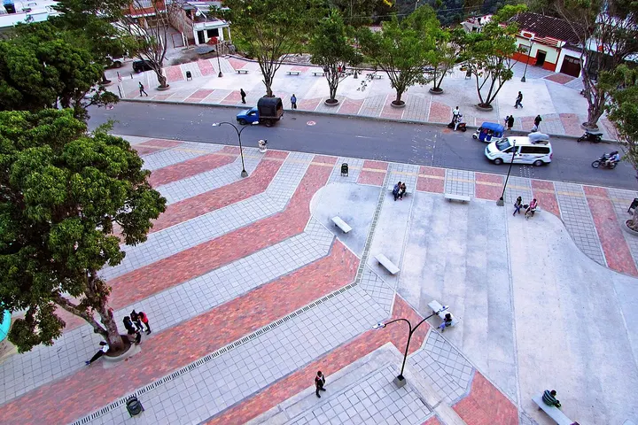 En primer plano, una plaza con árboles y bancos, con una calle y un vehículo en el fondo