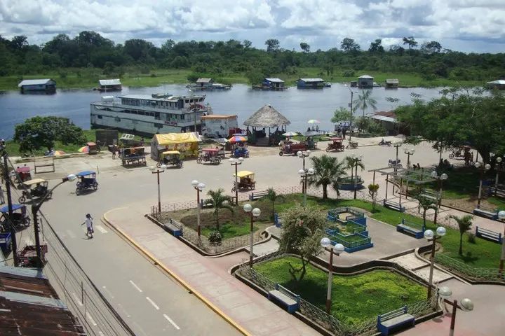 En primer plano, un parque con un lago, un camino y una plaza con árboles y bancos