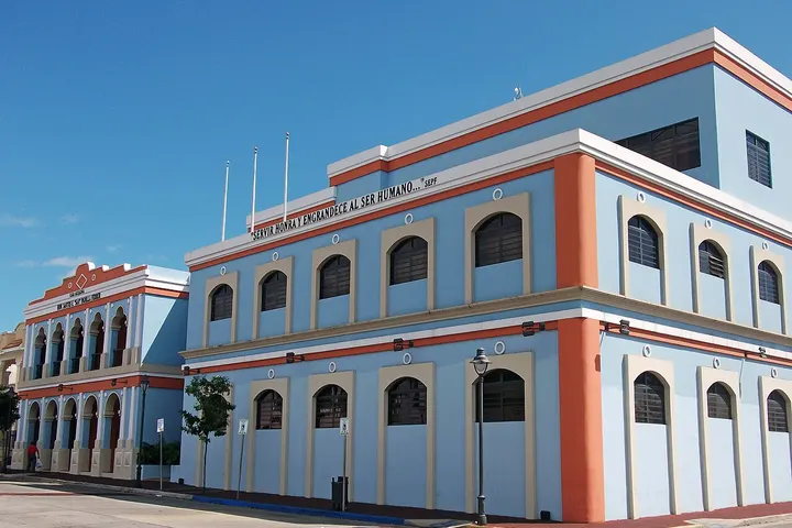 En primer plano, un edificio de color azul claro con detalles naranja y blanco, con arcos y ventanas