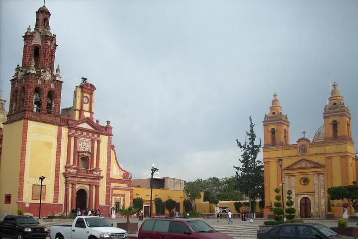 En primer plano, dos iglesias de colores amarillo y rojo, con torres y campanarios, rodeadas de coches
