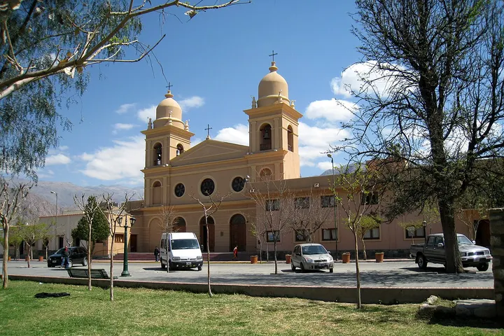 En primer plano, una iglesia con dos torres y una cúpula, rodeada de árboles y coches