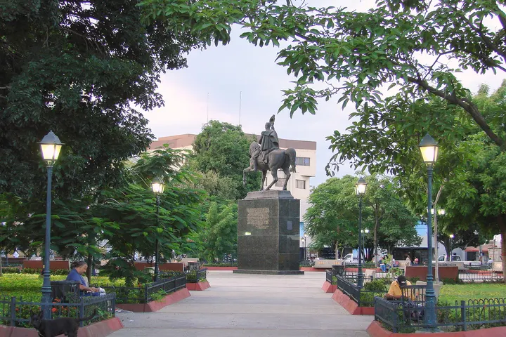 En primer plano, una estatua de caballo sobre un pedestal, rodeada de árboles y farolas