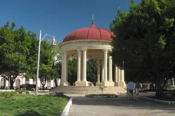 En primer plano, un kiosco con una cúpula roja y columnas blancas, rodeado de árboles y una zona verde