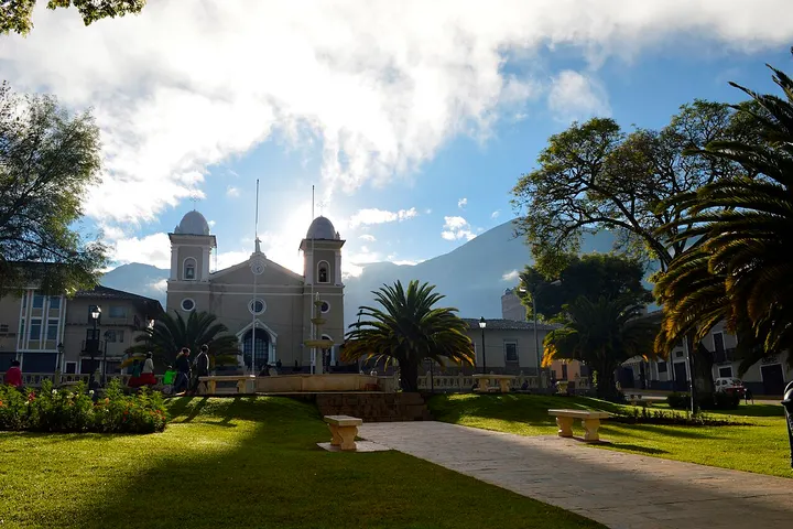 En primer plano, una iglesia blanca con dos torres, rodeada por un parque con árboles y césped