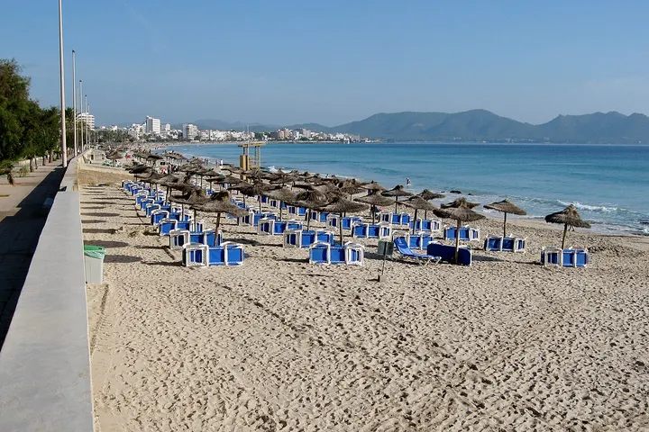 En primer plano, una playa con sillas de playa azules y sombrillas de paja