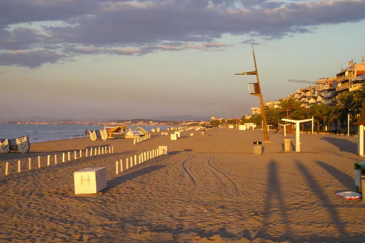 En primer plano, una playa con un par de huellas de neumáticos, y en la distancia, un puerto