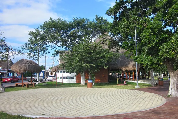 En primer plano, una plaza circular con una fuente en el centro, rodeada de árboles y una queja