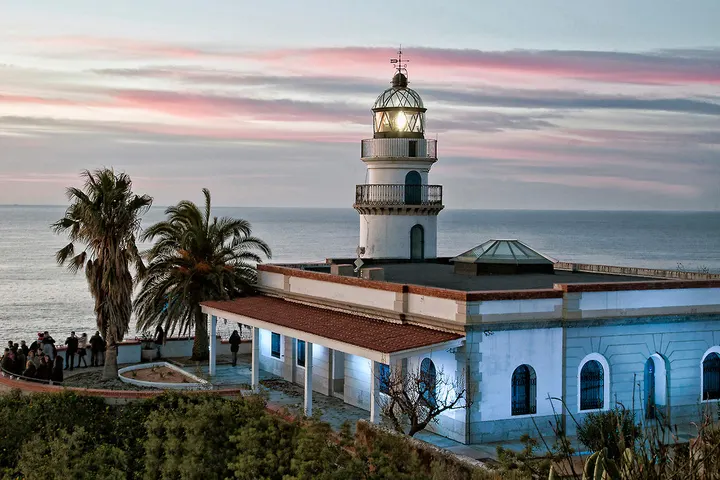 En primer plano, un faro blanco con una luz encendida, rodeado de palmeras y una casa azul