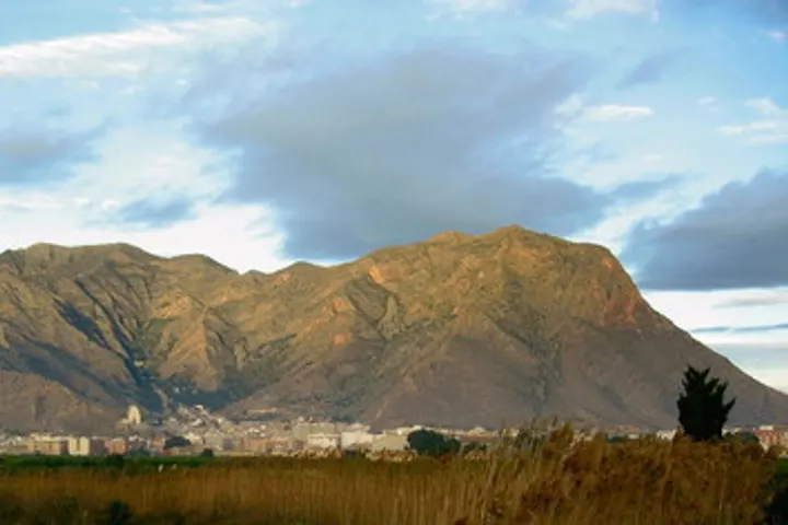 En primer plano, una montaña con un pico rocoso, y en segundo plano, una ciudad con edificios blancos