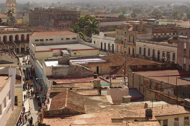 En primer plano, se ve una calle con gente, casas con tejados de tejas rojas y una torre de iglesia