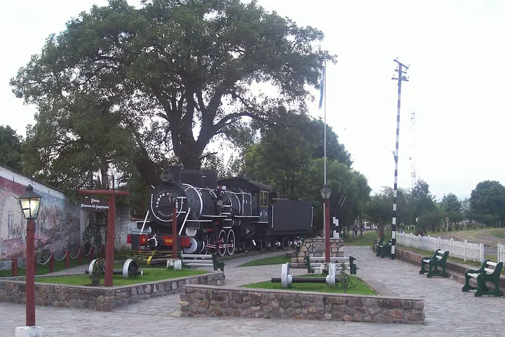 En primer plano, un tren antiguo de vapor está estacionado en un parque, rodeado de árboles y bancos