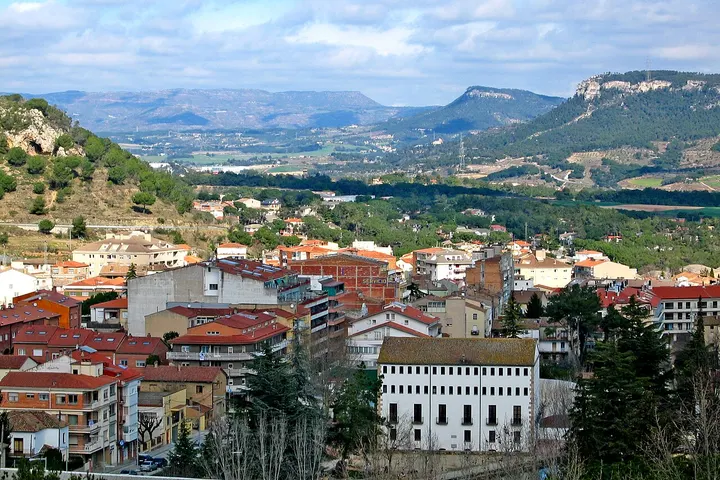 En primer plano, un edificio blanco con muchos ventanas, rodeado de árboles y con una vista panorámica de la montaña