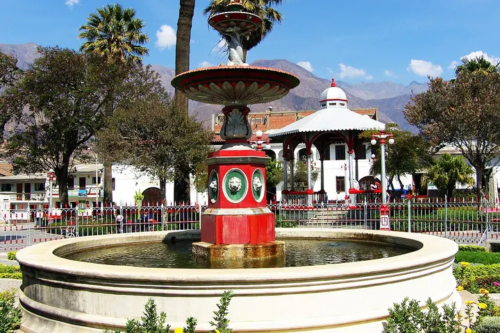 En primer plano, una fuente de color rojo y blanco, rodeada de un jardín con flores y árboles