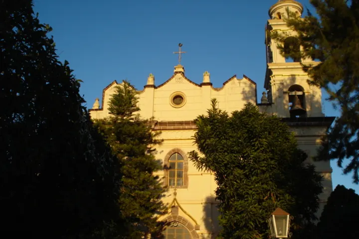 En primer plano, una iglesia blanca con una cruz en el techo, rodeada de árboles