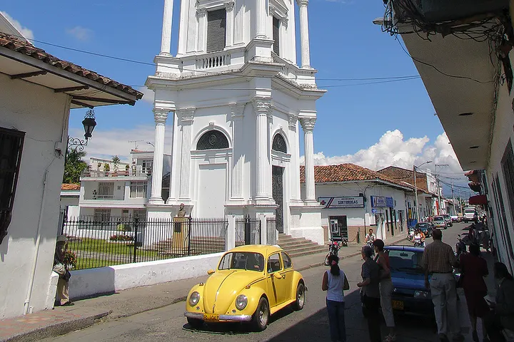 En primer plano, un Volkswagen Beetle amarillo, en una calle con edificios blancos y un cielo azul con nubes