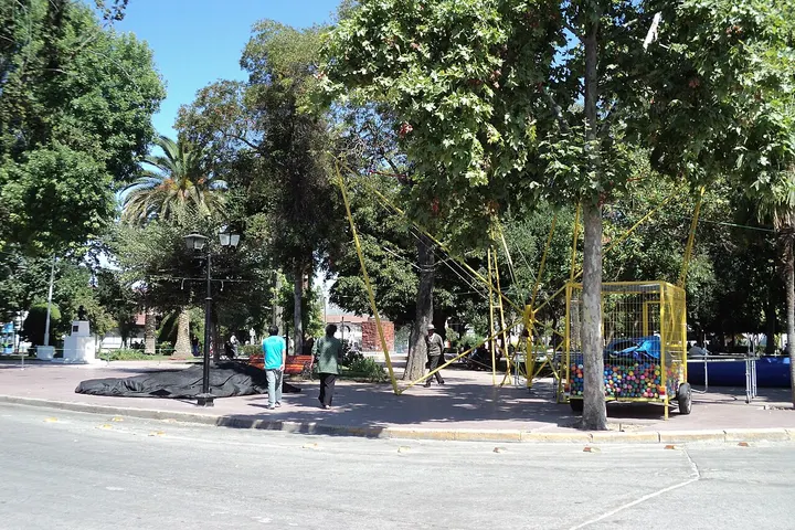 En primer plano, dos personas caminan por el parque, rodeadas de árboles y una estructura amarilla