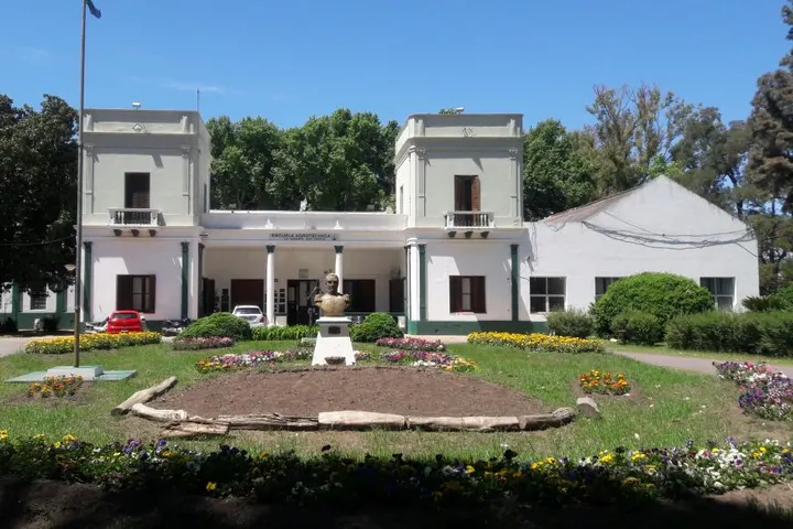 Edificio blanco de dos torres de una escuela agrotécnica con un busto y jardines en primer plano
