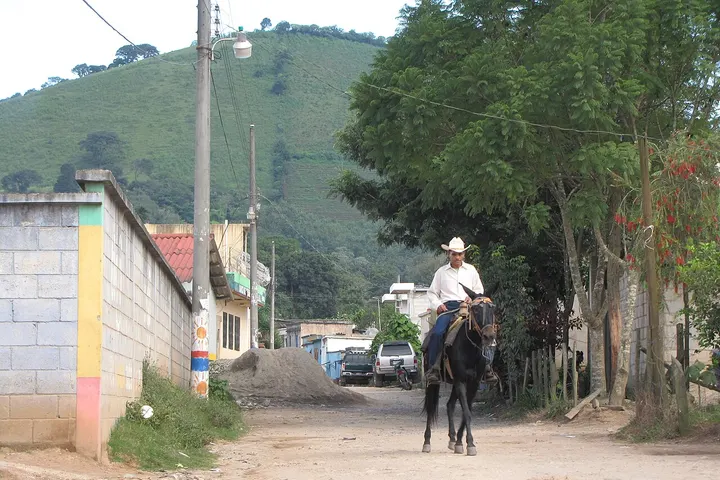 Un hombre monta a caballo por una calle en medio de una zona rural con casas y montañas al fondo